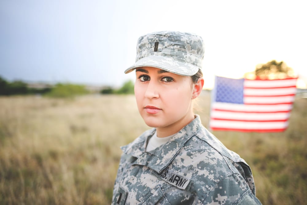 Service member in uniform standing outdoors with an American flag in the background.